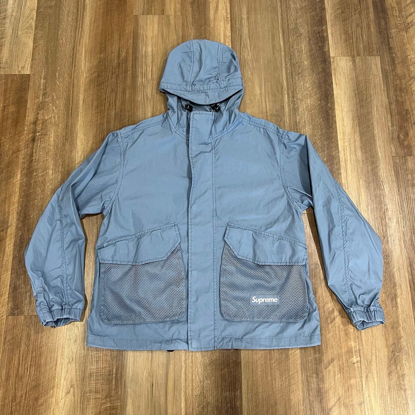 Laid flat on a wooden floor, the Supreme Mesh Pocket Cargo Jacket in Slate exudes a casual style. This 100% authentic preowned piece, featuring long sleeves, combines comfort and flair.