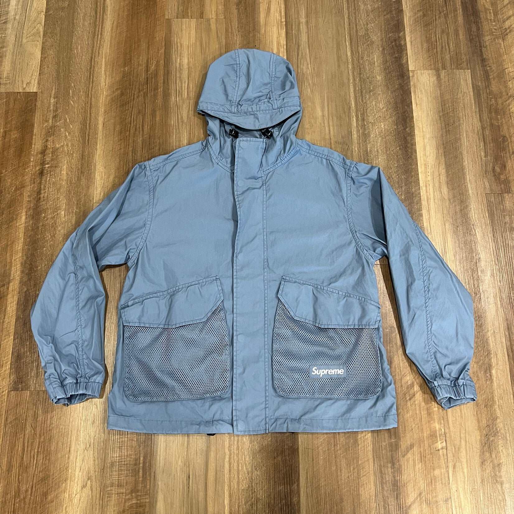 Laid flat on a wooden floor, the Supreme Mesh Pocket Cargo Jacket in Slate exudes a casual style. This 100% authentic preowned piece, featuring long sleeves, combines comfort and flair.