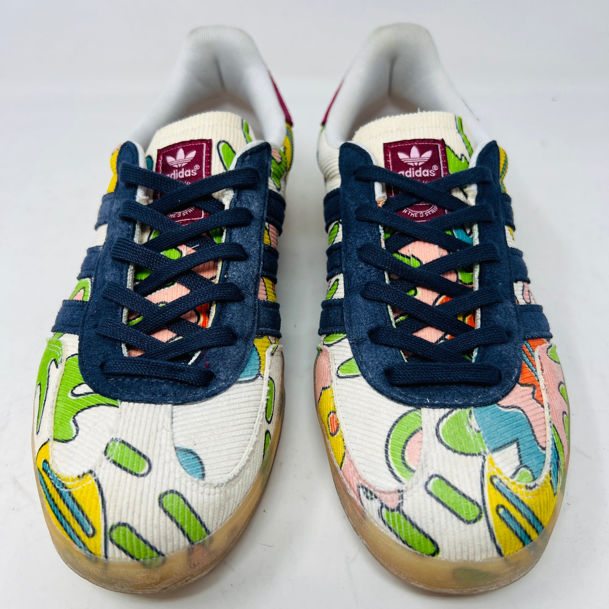 The Adidas Gazelle Indoor Sean Wotherspoon sneaker, with colorful abstract uppers in yellow, red, pink, and green, dark blue stripes, red laces, and translucent soles, rests on a blue box showing size and product details.