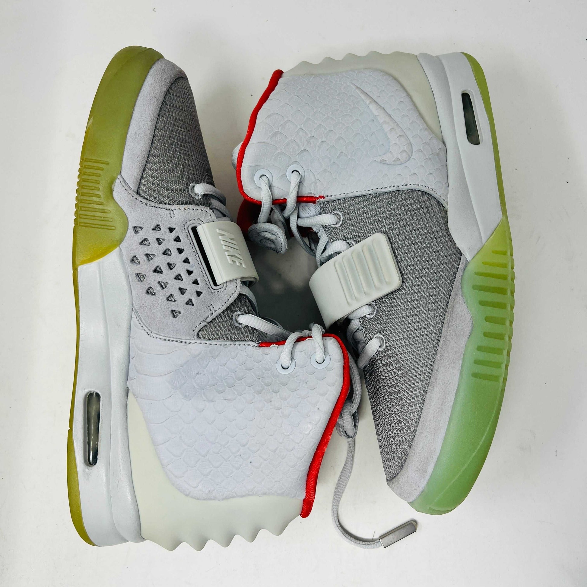 A pair of new Nike Air Yeezy 2 Pure Platinum high-tops rests on a cardboard box. The sneakers are mostly gray with a red lining, midfoot strap, and green-tinted soles. The label reads Air Yeezy 2 NRG, size 9, showcasing authentic detailing by Nike.