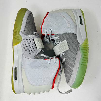 A pair of new Nike Air Yeezy 2 Pure Platinum high-tops rests on a cardboard box. The sneakers are mostly gray with a red lining, midfoot strap, and green-tinted soles. The label reads Air Yeezy 2 NRG, size 9, showcasing authentic detailing by Nike.