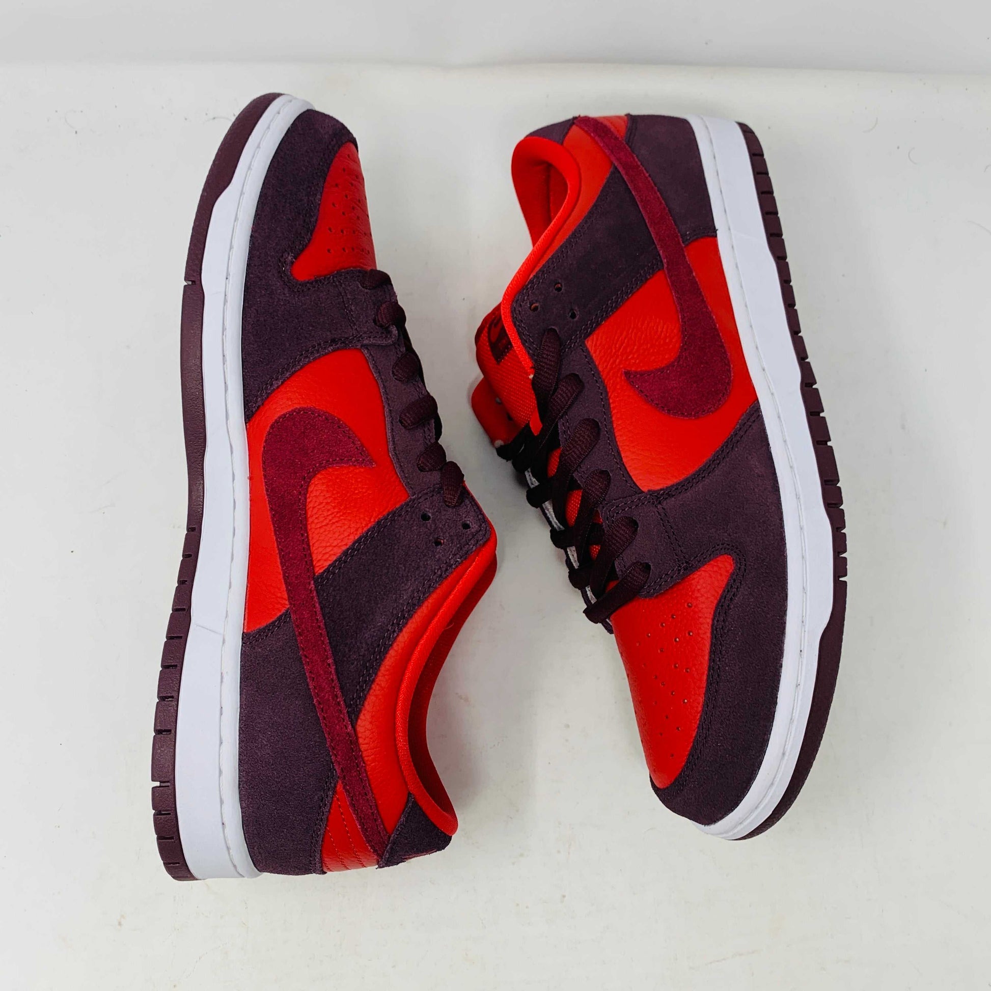 A 100% authentic, brand new Nike SB Dunk Low Cherry sneaker in red and dark brown sits on a branded purple Nike SB shoebox. It features a red swoosh, toe box, dark brown overlays, extra laces, white midsole, and dark brown outsole.