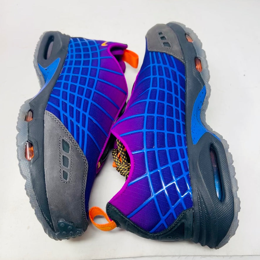Displayed on an orange Nike shoe box is the colorful Nike Air Max Sunder SP Kids of Immigrants Sun Down sneaker, featuring a grid pattern with shades of purple, blue, and gray, complemented by orange accents and a swoosh logo. The shoe box shows a k-i logo inside a circle.