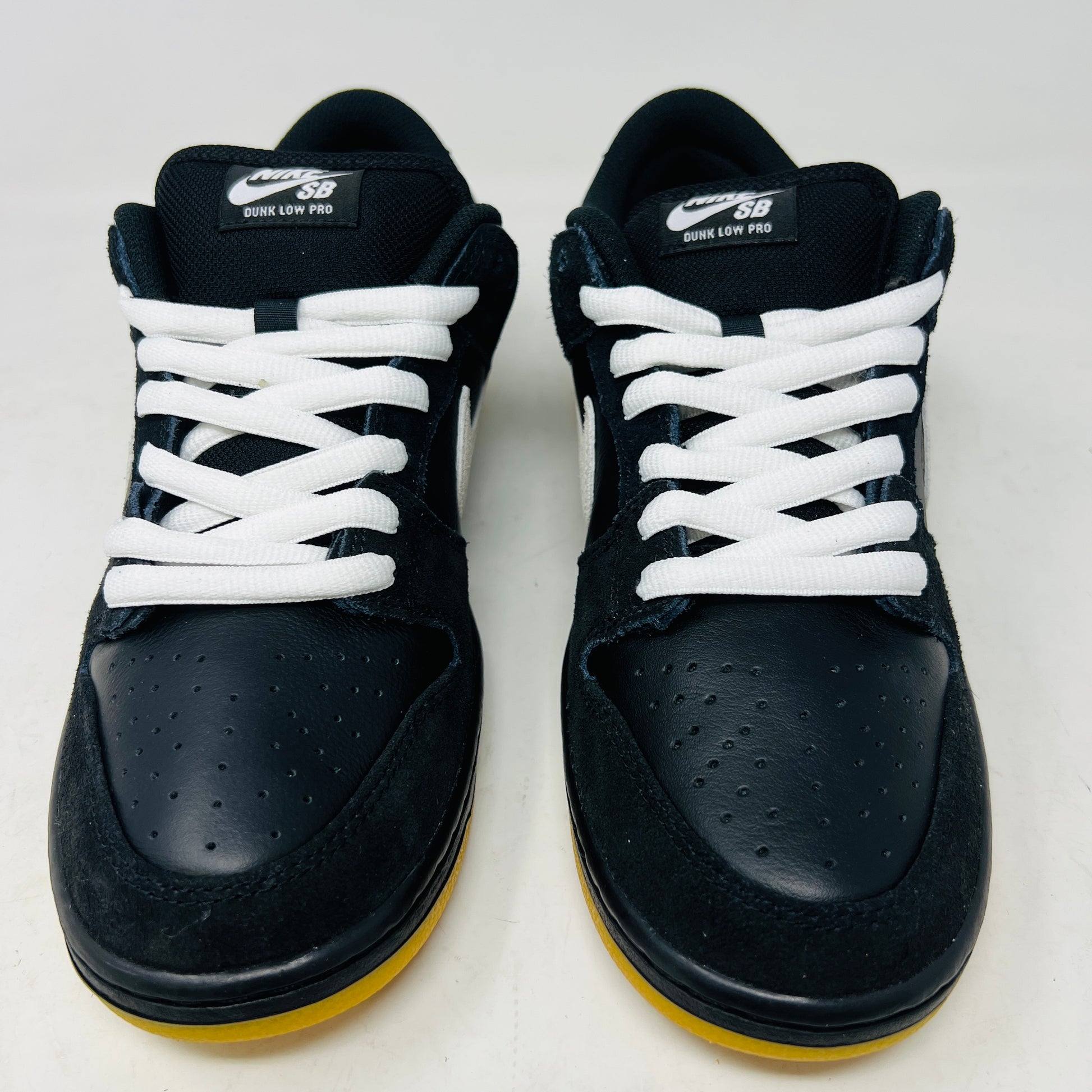A black Nike Dunk Low Pro SB Black White Gum sneaker with white laces and swoosh sits on a purple Nike SB box. The 2025 shoe has a yellow sole, leather-texture finish, and both are in new condition.