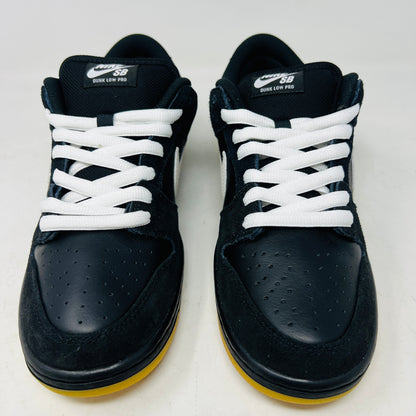 A black Nike Dunk Low Pro SB Black White Gum sneaker with white laces and swoosh sits on a purple Nike SB box. The 2025 shoe has a yellow sole, leather-texture finish, and both are in new condition.