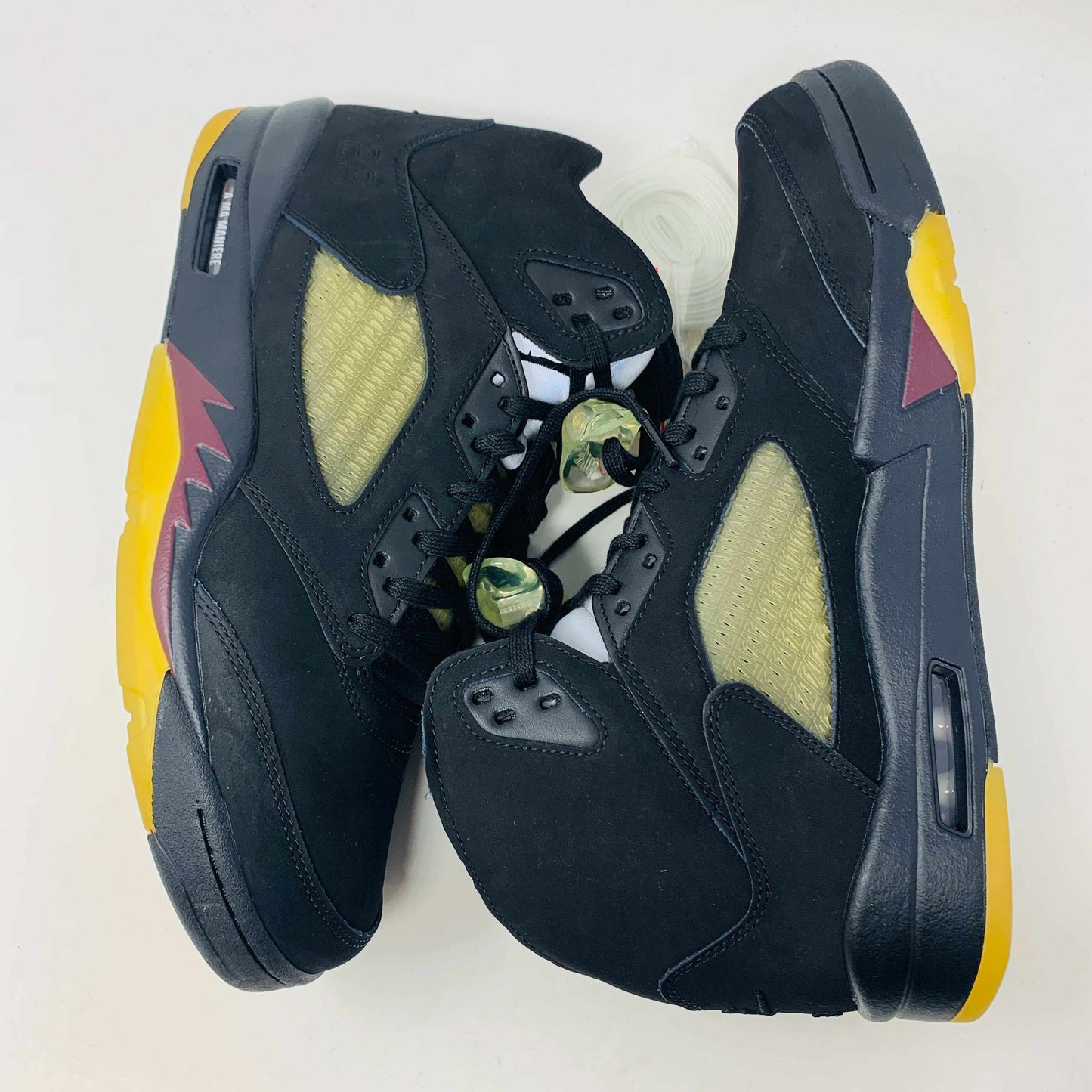 The Jordan 5 A Ma Maniére Dusk sneakers, by Jordan, feature a high-top black design with yellow and red accents, camo-patterned shoelace locks, and translucent netting on the sides, set against a plain background.