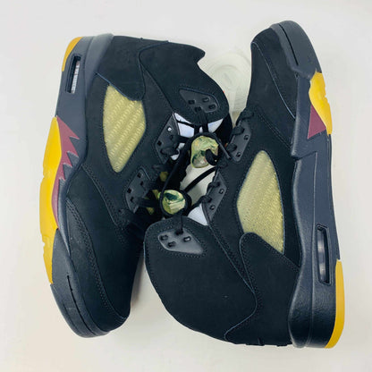 The Jordan 5 A Ma Maniére Dusk sneakers, by Jordan, feature a high-top black design with yellow and red accents, camo-patterned shoelace locks, and translucent netting on the sides, set against a plain background.