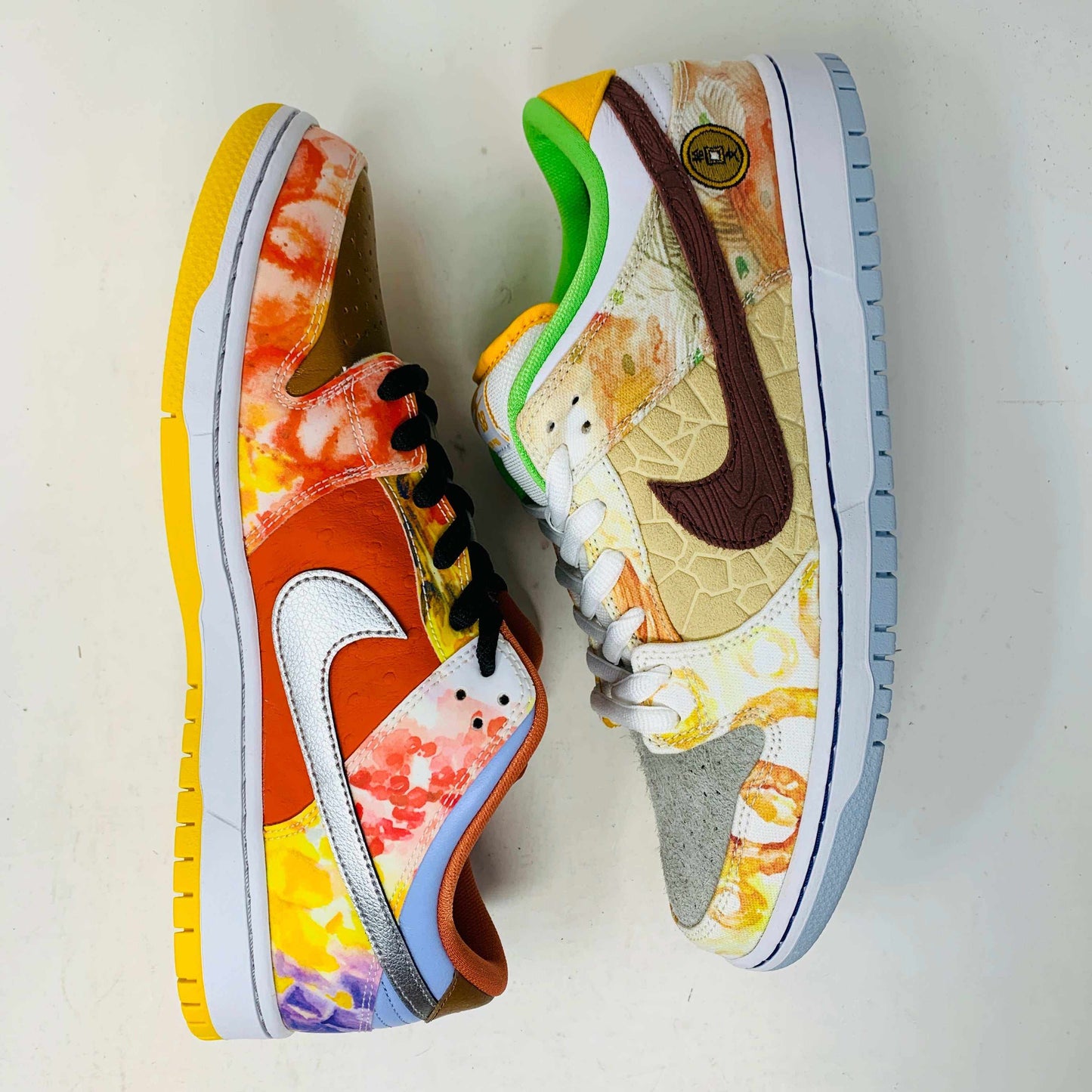 The Nike SB Dunk Low Street Hawker is a colorful sneaker displayed on a green and yellow box, featuring an authentic artistic design with brown, green, and orange tones. It has a quilted texture, white swoosh, white sole with light gray bottom, and comes with extra laces.