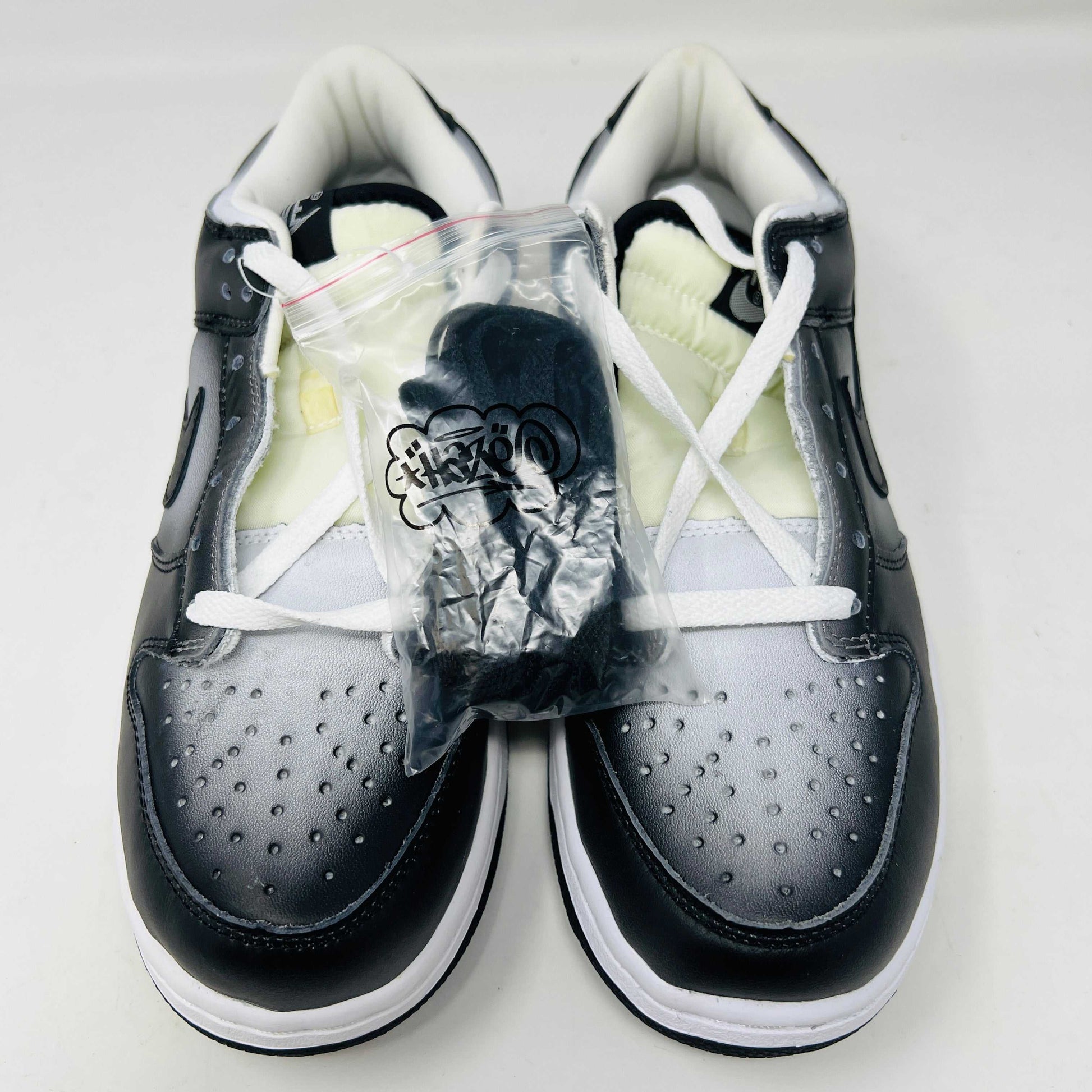 Authentic Nike Dunk Low Haze sneakers in black and white with white laces are displayed front-on against a white backdrop. A clear bag with extra black laces is on the left shoe.