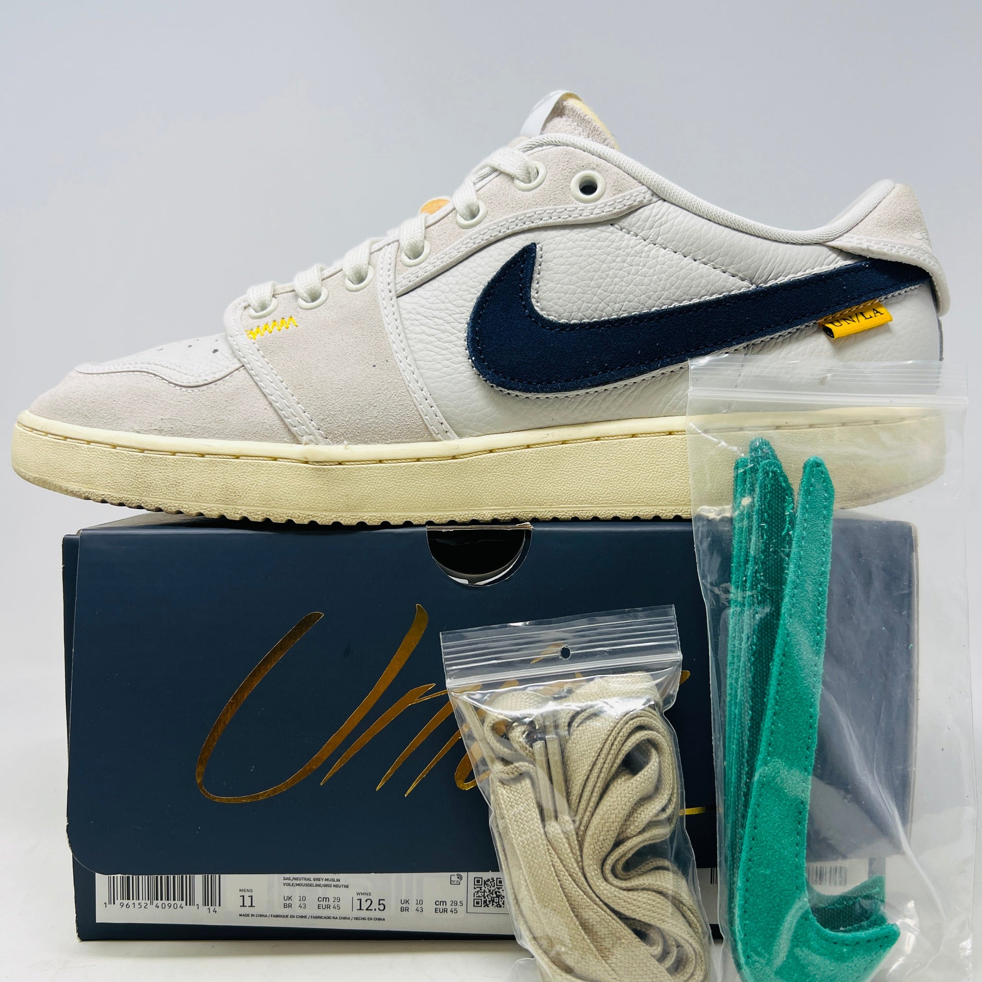 The Jordan 1 Retro AJKO Low SP Union Sail Leather features a beige and navy design with a navy Swoosh, a vintage-style sole, and arrives on a shoebox. Includes extra laces and green inserts in separate bags.