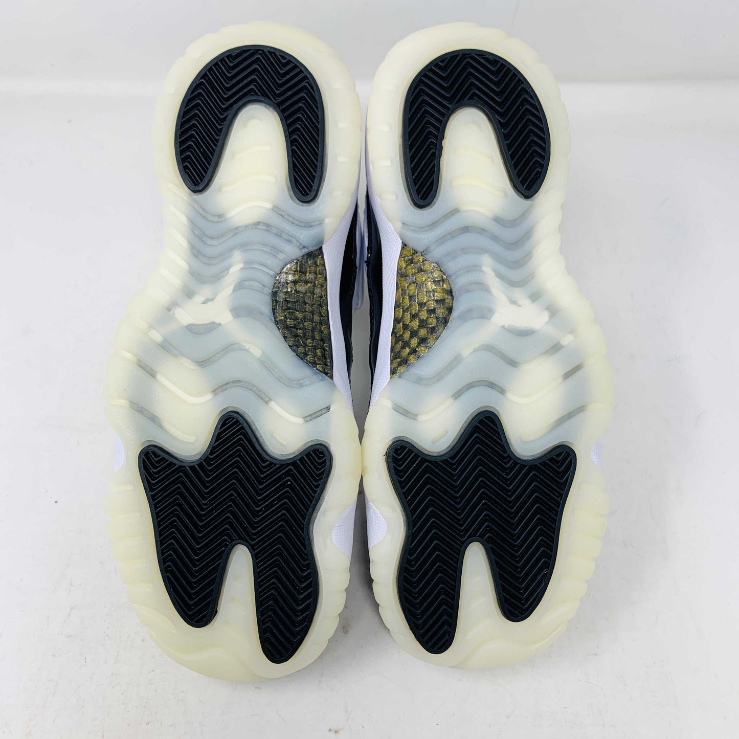 A pair of authentic Jordan 11 Retro DMP Gratitude (2023) (GS) sneakers, featuring a glossy white and black finish with a gold logo, is placed on top of a gray Nike Air shoebox.