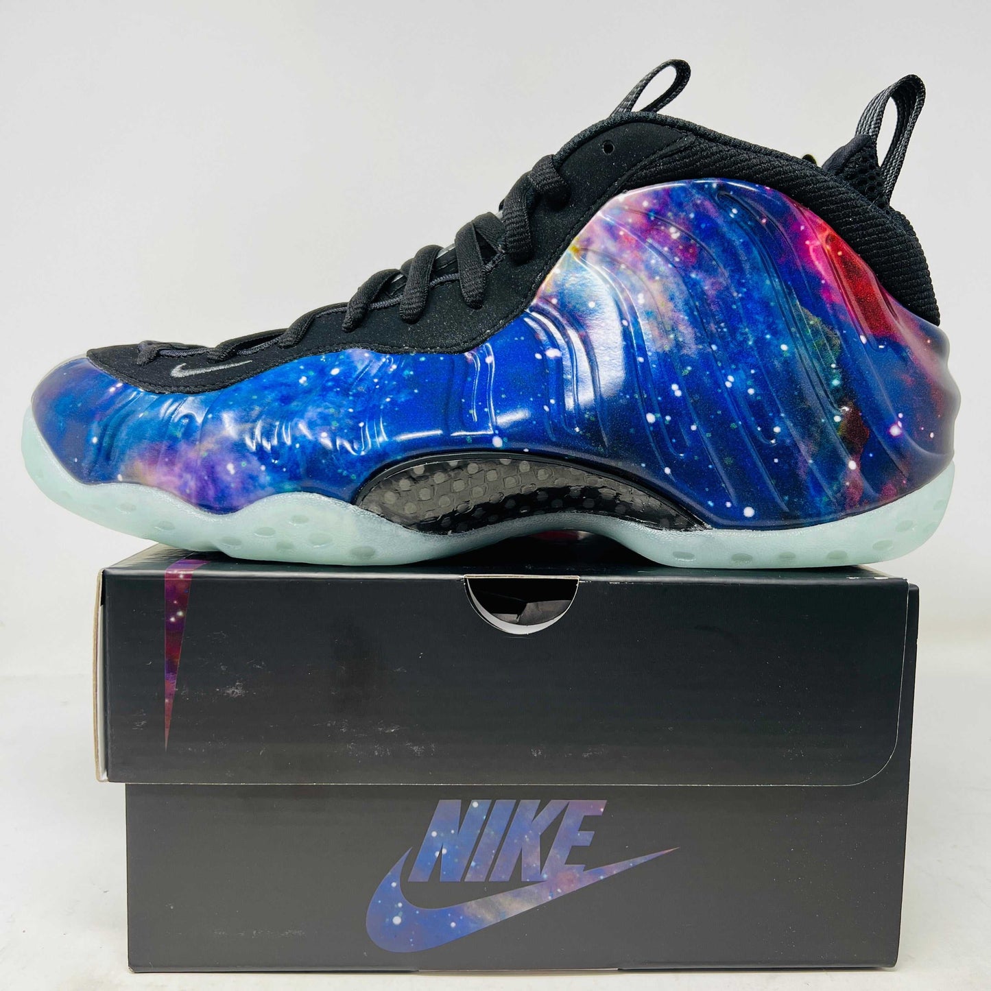 The 2025 Nike Air Foamposite One Galaxy sneaker features a vibrant galaxy design on the upper, complemented by a dark sole and black laces. Presented atop its black box, this edition is an essential piece for collectors.