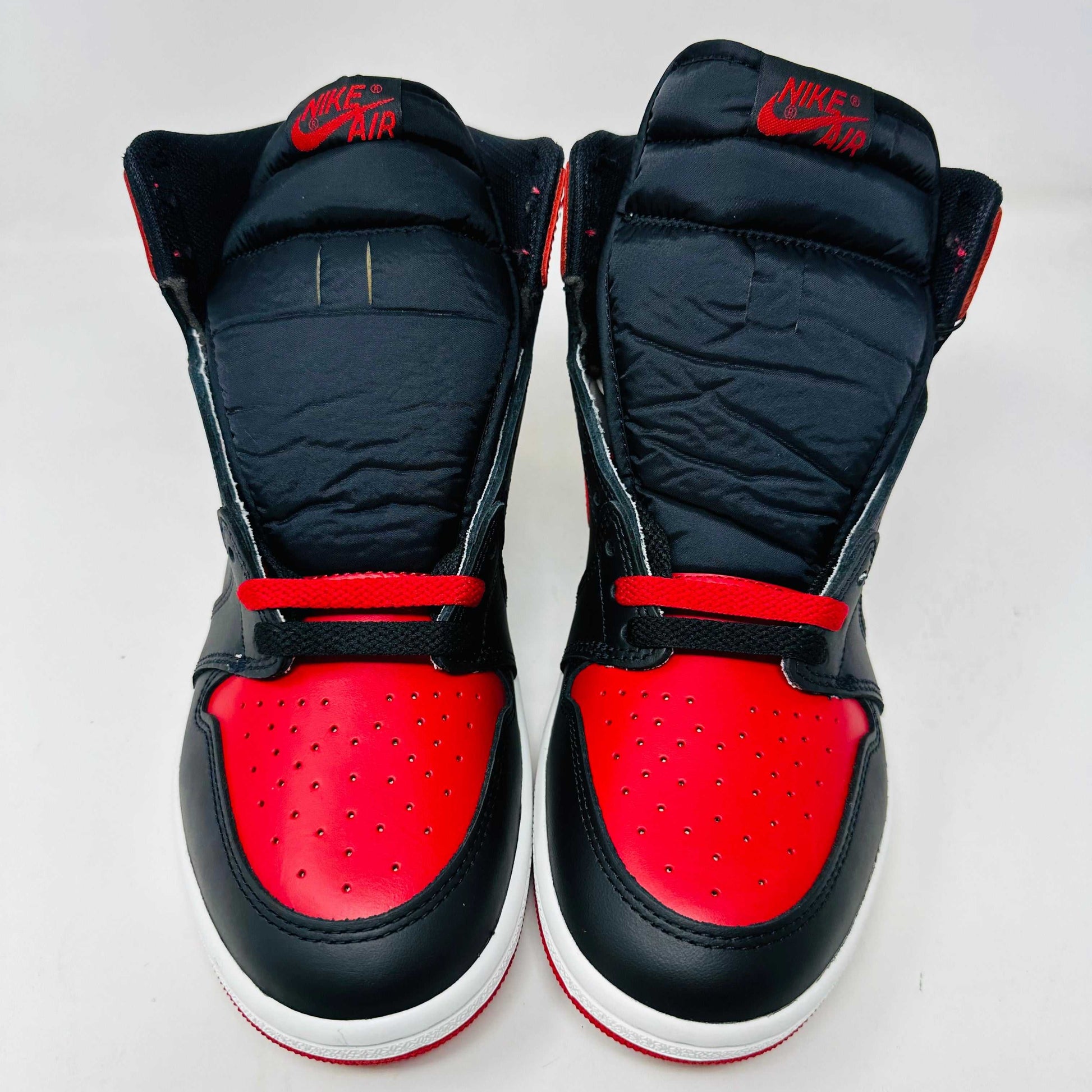 A Jordan 1 Retro High 85 OG Bred (2025) sneaker by Jordan sits on its matching shoebox, showcasing the iconic swoosh and Air Jordan logo. Its styled in black and red with a white midsole, bold red laces, and detailed black accents.