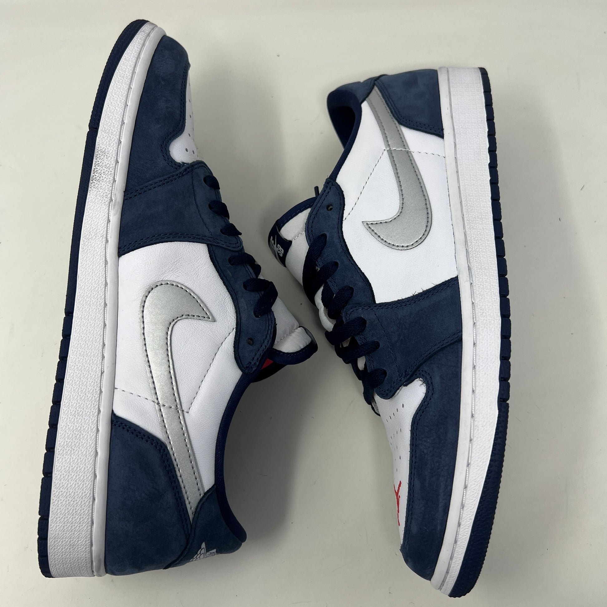 A Jordan 1 Low SB Midnight Navy shoe from Jordan, featuring blue toe and heel, white side panels, and a silver swoosh, is displayed on its box. The VERY CLEAN UPPERS are complemented by the size and product info visible on the label.