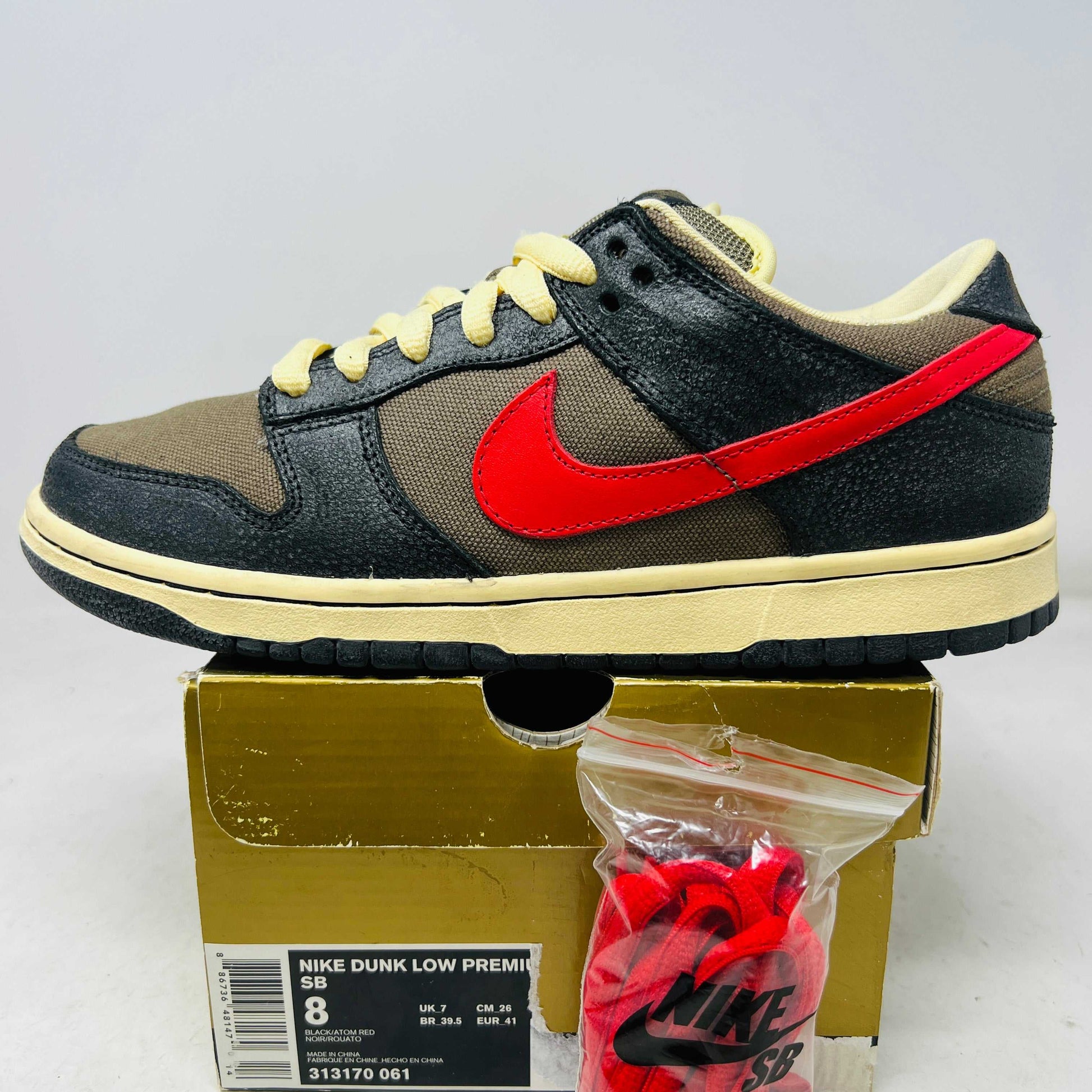 Nike SB Dunk Low Iron Man sneaker with black and brown upper, red Nike swoosh, cream laces, and additional red laces in a bag on a gold Nike box