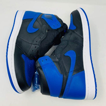 A pair of brand new black and blue Jordan 1 Royal 2017 high-tops are showcased on a black and red Nike box, with a small plastic bag holding authentic blue laces attached to one shoe.