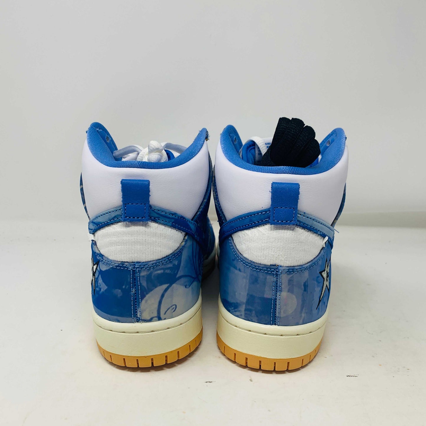 The Nike SB Dunk High Carpet Company sneakers feature a blue and white starry design, presented on a green and yellow Nike shoe box. Three extra laces in blue, yellow, and white are included to ensure your style is 100% authentic.