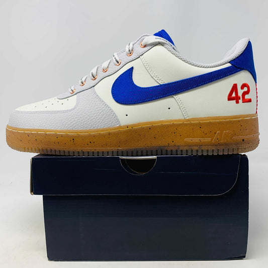 The Nike Air Force 1 Low Jackie Robinson sneaker features a white upper with a blue Nike swoosh, blue heel tab, orange eyelets, white laces, and brown speckled sole. It sits atop a black box with an authentic red 42 on the heel.