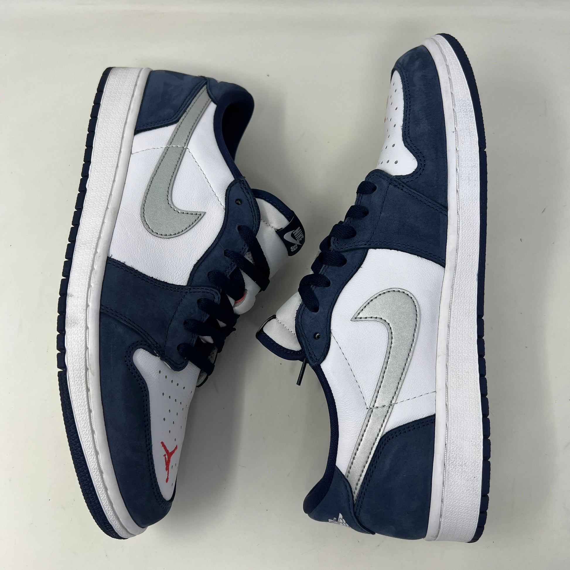 A Jordan 1 Low SB Midnight Navy shoe from Jordan, featuring blue toe and heel, white side panels, and a silver swoosh, is displayed on its box. The VERY CLEAN UPPERS are complemented by the size and product info visible on the label.