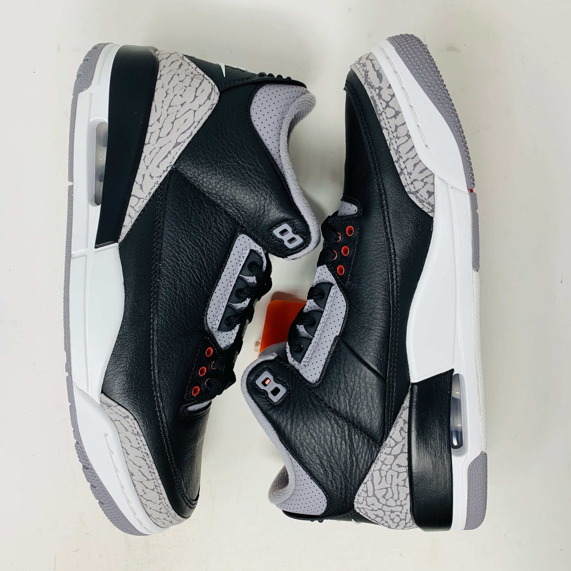 The Jordan 3 Retro OG Black Cement (2024) sneaker, featuring black and gray tones with red accents, is displayed on a sleek black shoebox. With textured details and visible air cushioning, an orange Nike Air tag authenticates this stylish addition to your collection.