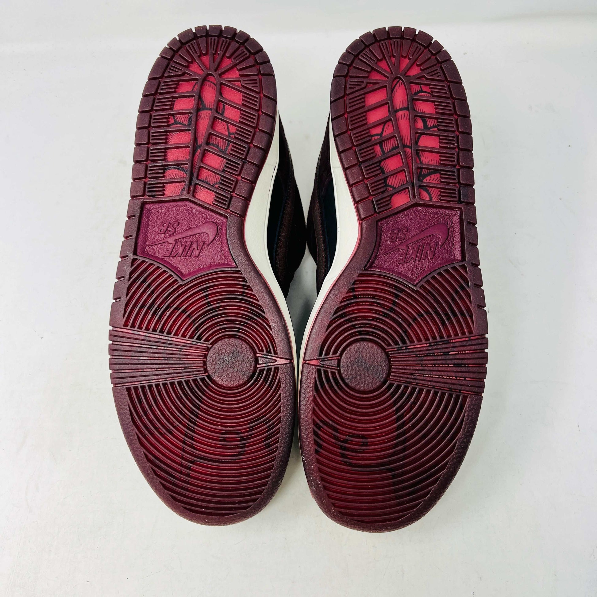 A BRAND NEW Nike SB Dunk Low Riot Skateshop sneaker featuring a black design with maroon accents, including a swoosh, laces, and a black sole with a white midsole, is displayed on top of its original white Nike SB shoebox.