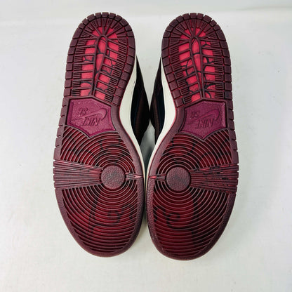A BRAND NEW Nike SB Dunk Low Riot Skateshop sneaker featuring a black design with maroon accents, including a swoosh, laces, and a black sole with a white midsole, is displayed on top of its original white Nike SB shoebox.