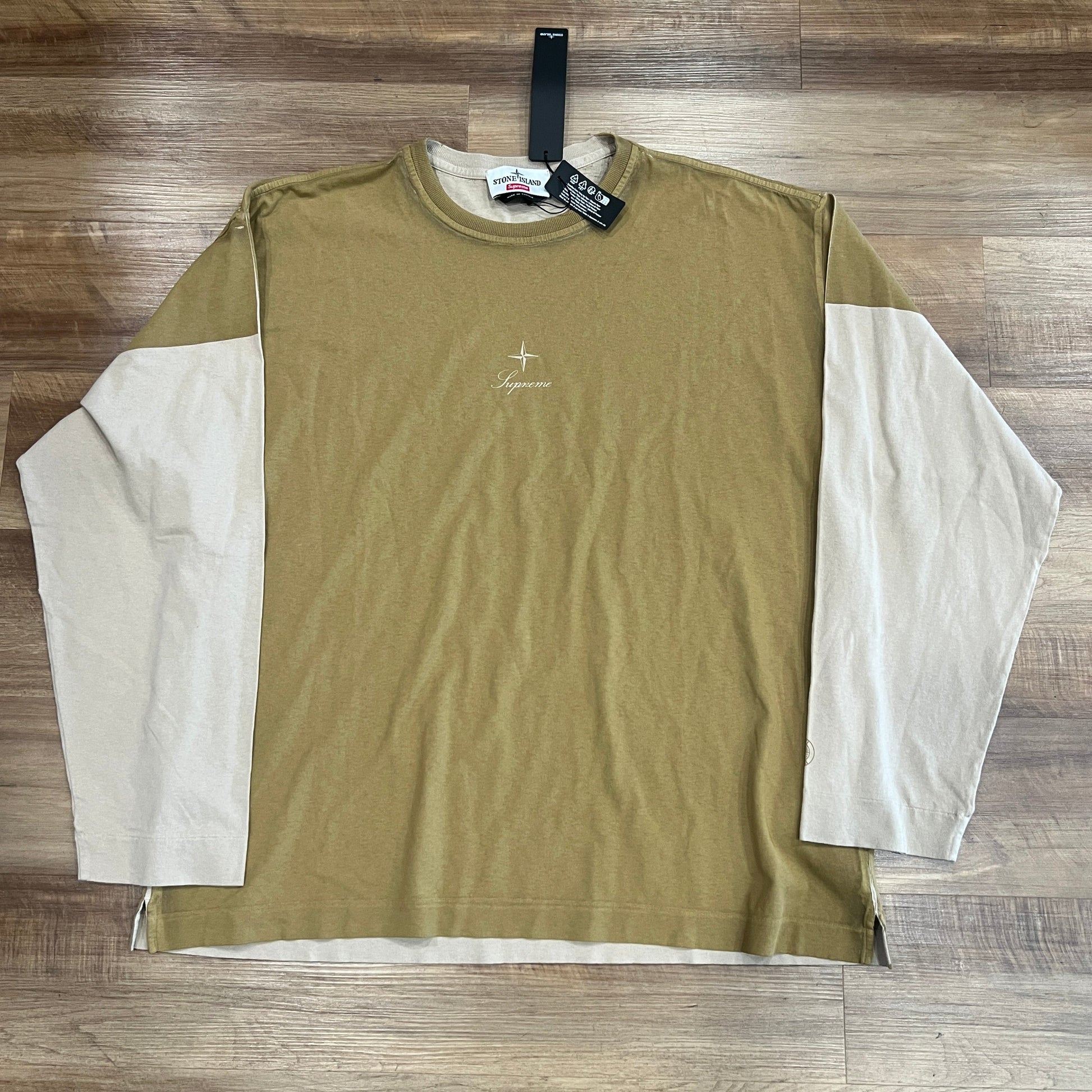 A brand new Supreme Stone Island L/S Top in tan, size large, is laid flat on a wooden floor. It features beige sleeves, a small logo on the chest, and a black tag attached to the collar.