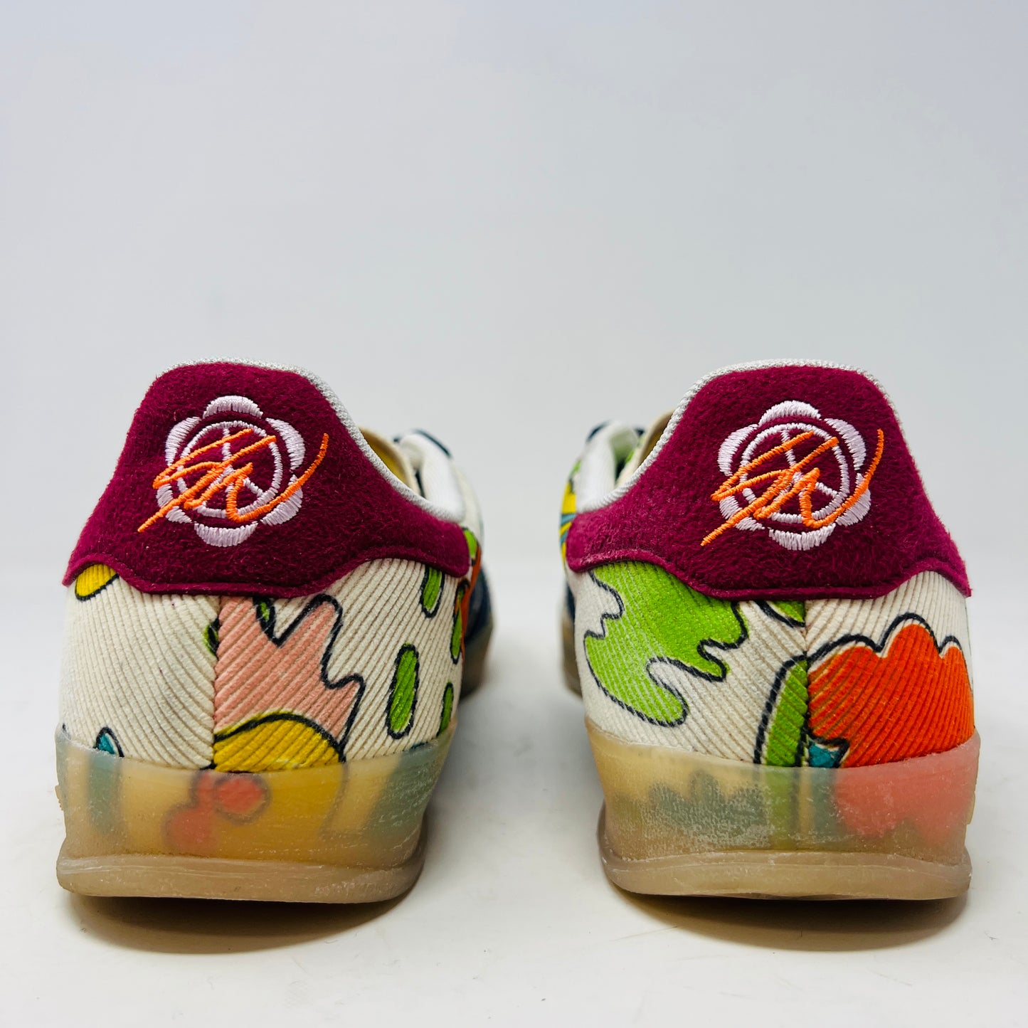 The Adidas Gazelle Indoor Sean Wotherspoon sneaker, with colorful abstract uppers in yellow, red, pink, and green, dark blue stripes, red laces, and translucent soles, rests on a blue box showing size and product details.