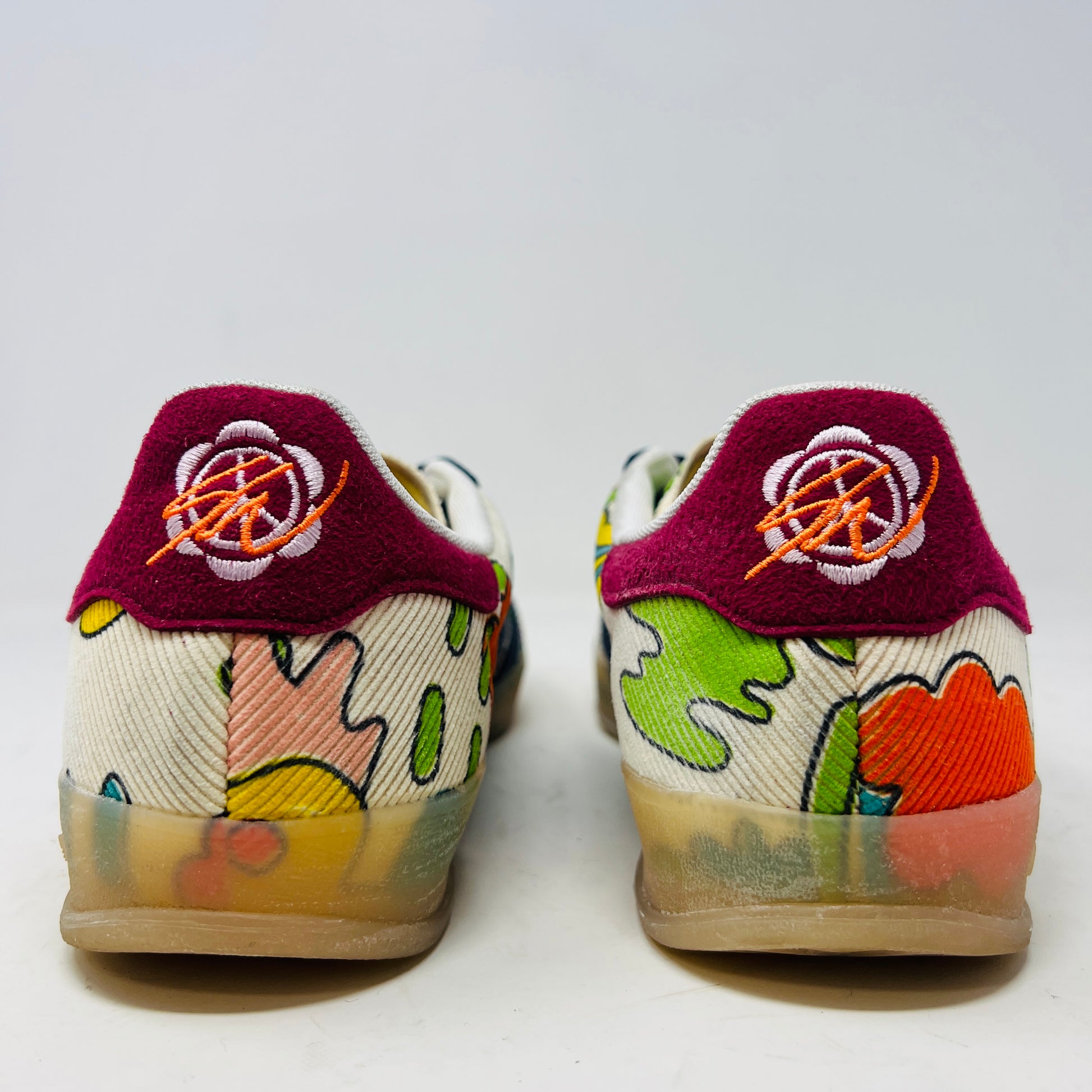 The Adidas Gazelle Indoor Sean Wotherspoon sneaker, with colorful abstract uppers in yellow, red, pink, and green, dark blue stripes, red laces, and translucent soles, rests on a blue box showing size and product details.