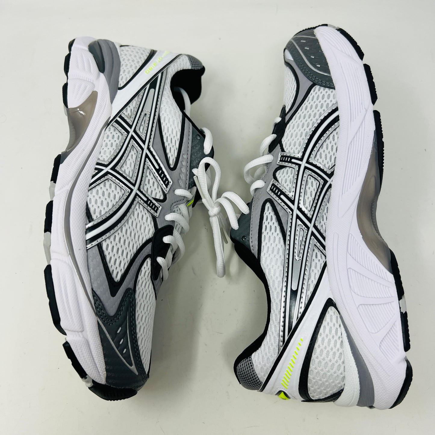 A brand new pair of ASICS GT-2160 JJJJound White sneakers, featuring mesh details and black stripes, is displayed on a black ASICS shoe box with extra laces in front for a complete authentic look.