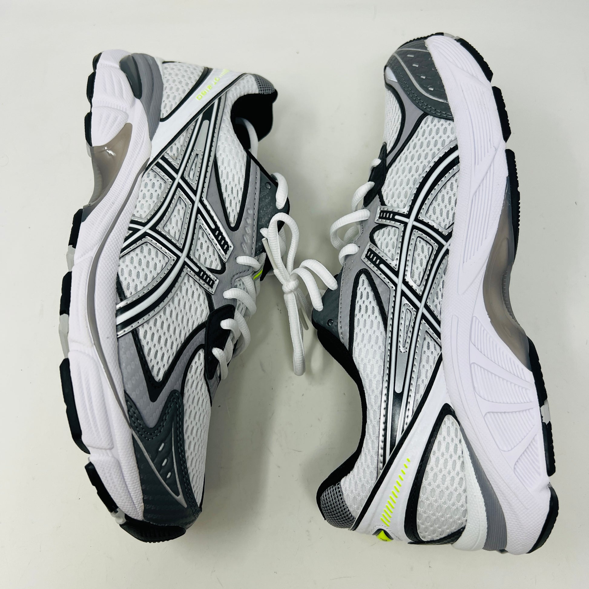 A brand new pair of ASICS GT-2160 JJJJound White sneakers, featuring mesh details and black stripes, is displayed on a black ASICS shoe box with extra laces in front for a complete authentic look.