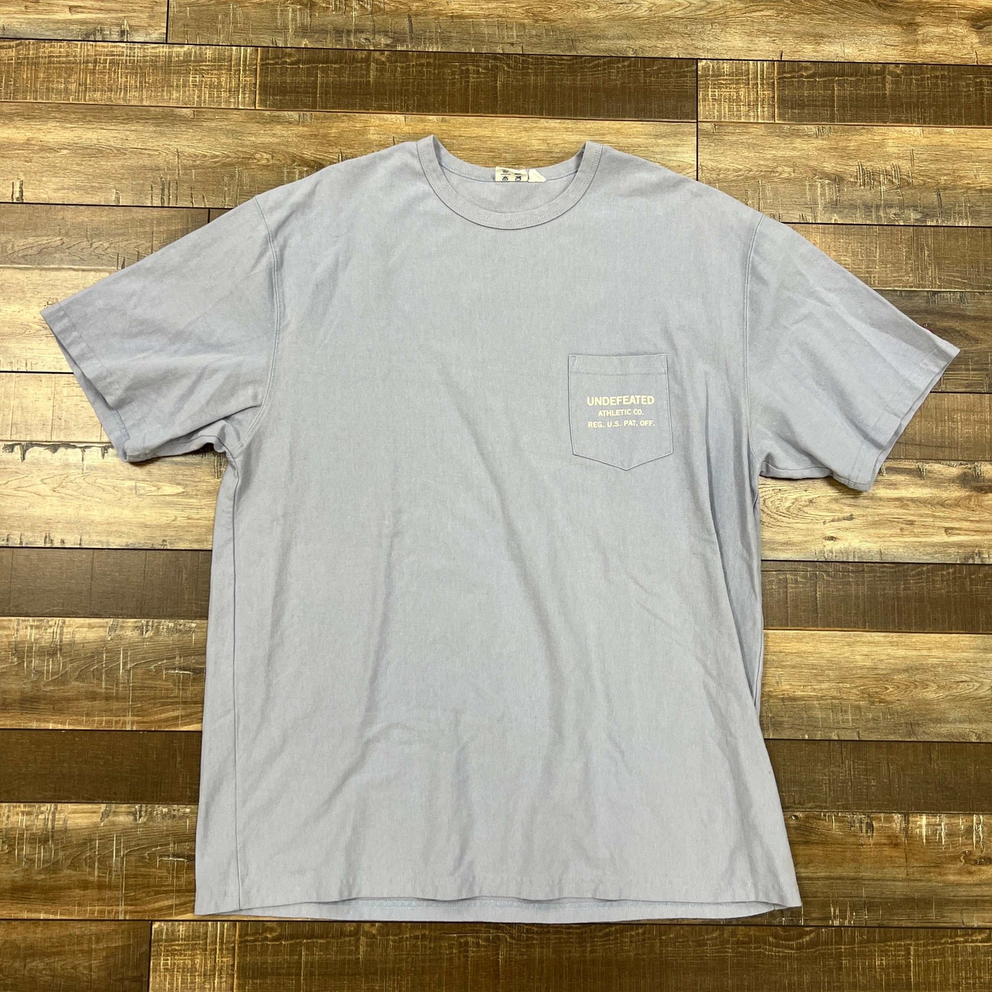 Undefeated Blue pocket tee shirt laid flat on wooden surface, preowned condition