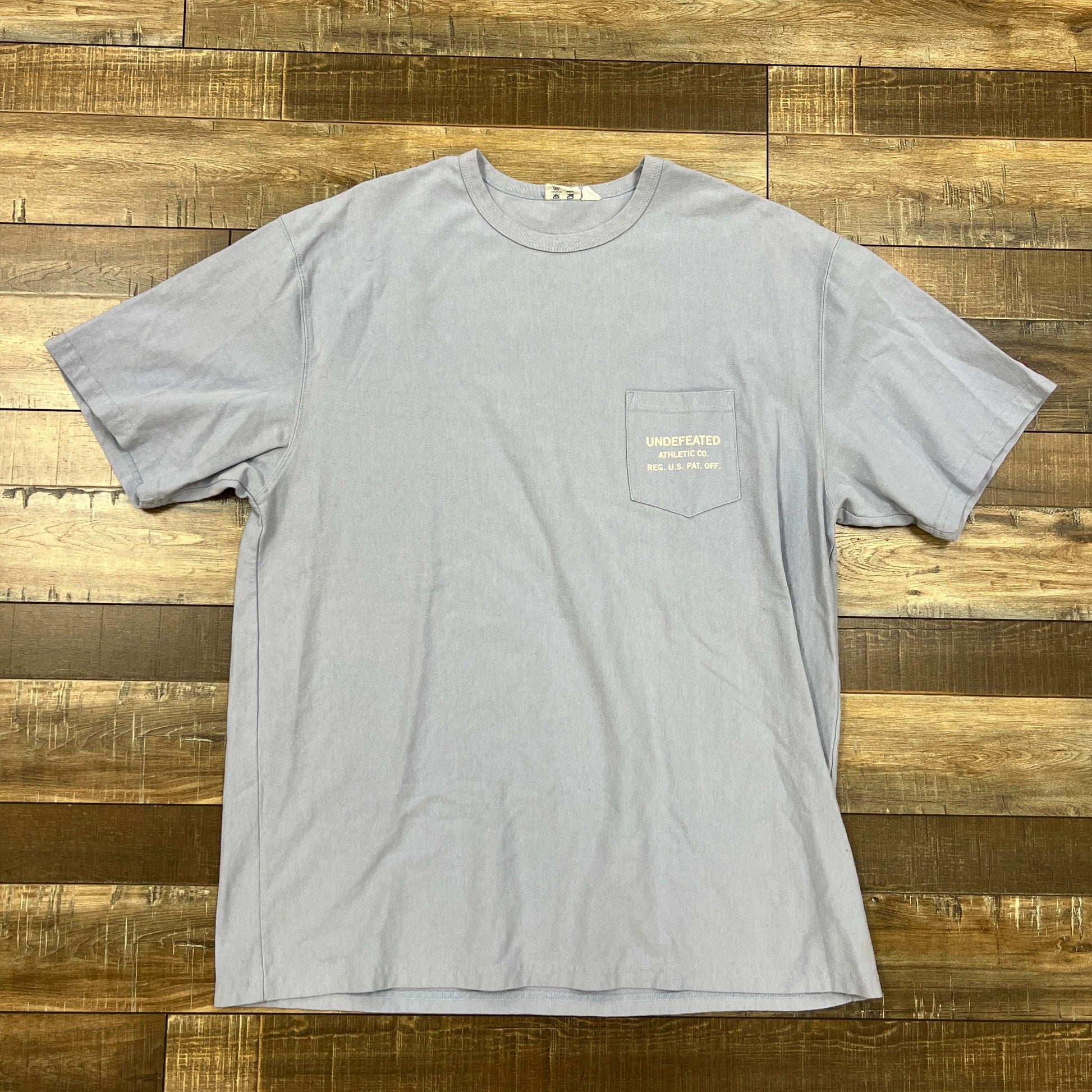 Undefeated Blue pocket tee shirt laid flat on wooden surface, preowned condition