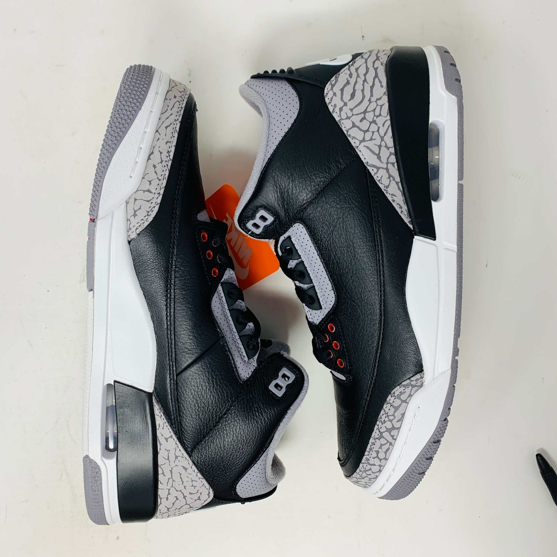 The Jordan 3 Retro OG Black Cement (2024) sneaker, featuring black and gray tones with red accents, is displayed on a sleek black shoebox. With textured details and visible air cushioning, an orange Nike Air tag authenticates this stylish addition to your collection.