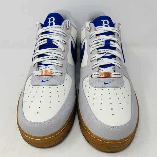 The Nike Air Force 1 Low Jackie Robinson sneaker features a white upper with a blue Nike swoosh, blue heel tab, orange eyelets, white laces, and brown speckled sole. It sits atop a black box with an authentic red 42 on the heel.