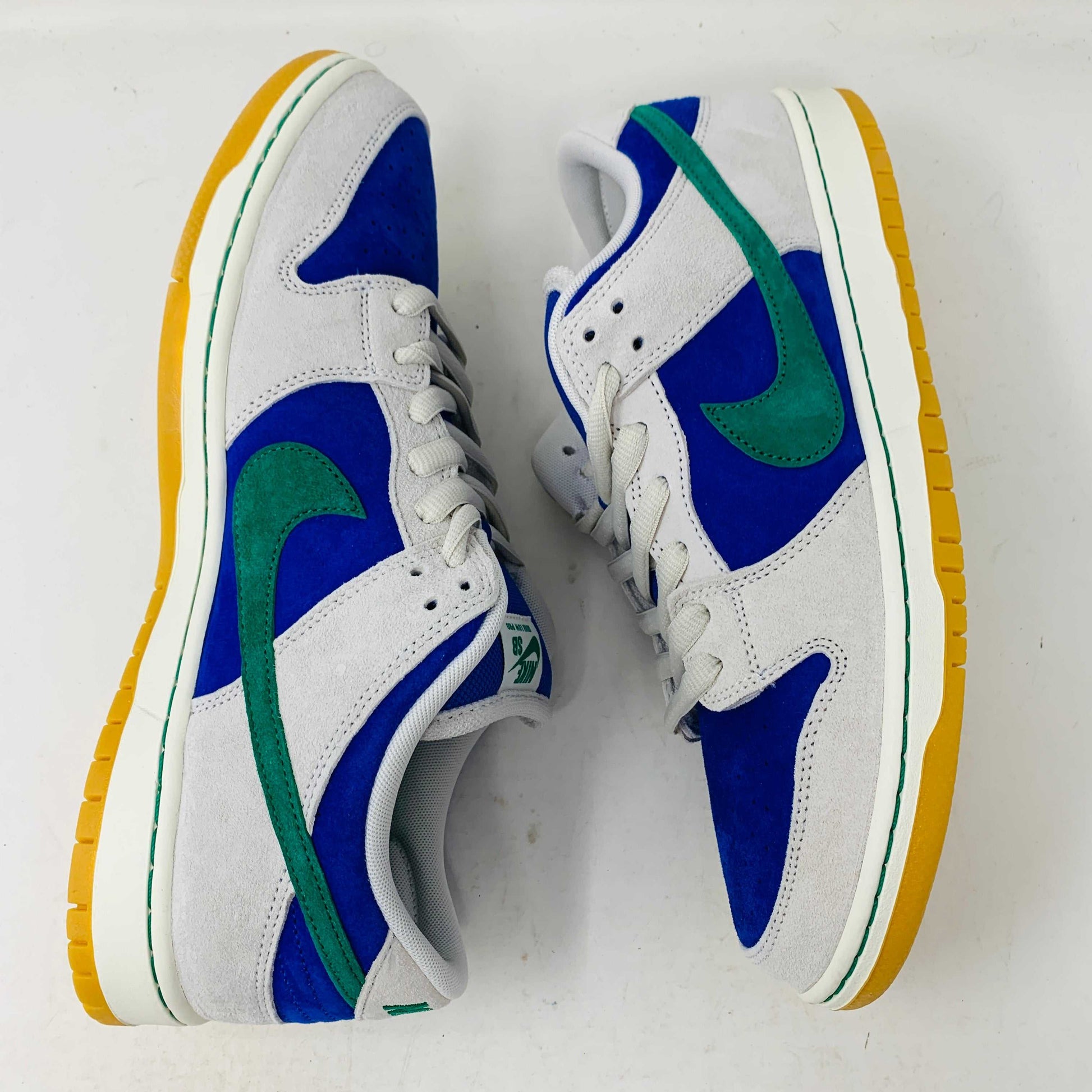 The Nike SB Dunk Low Hyper Royal Malachite, a fresh pair of white and blue sneakers with a green swoosh and gum sole, includes extra laces and is presented atop a purple Nike SB shoe box.