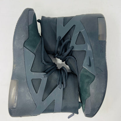 The Nike Air Fear of God 1 Triple Black is a high-top, dark, minimalist sneaker with angular overlays and a textured sole. It features a high collar, matte finish, dark laces, and has a small logo near the midsole. The background is light gray with general wear signs.