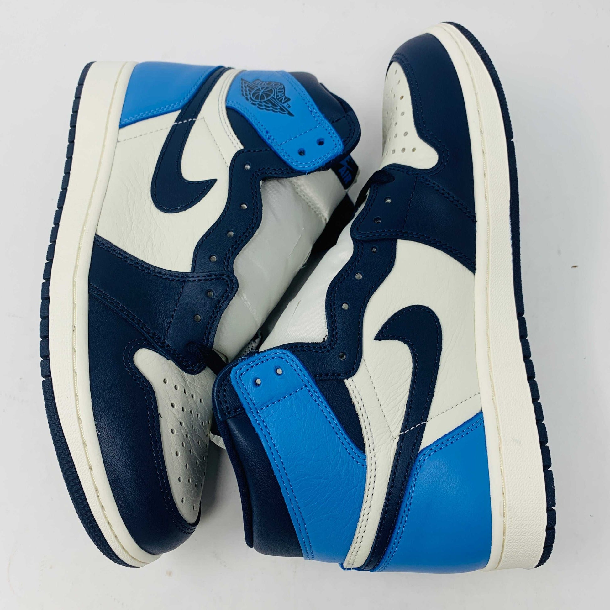 The new Jordan 1 Obsidian high-top sneaker, in blue and white with a black swoosh and blue laces, is displayed on a black and red box featuring 1985 Air Jordan 1 text. A printed Air Jordan wings logo is near the ankle.