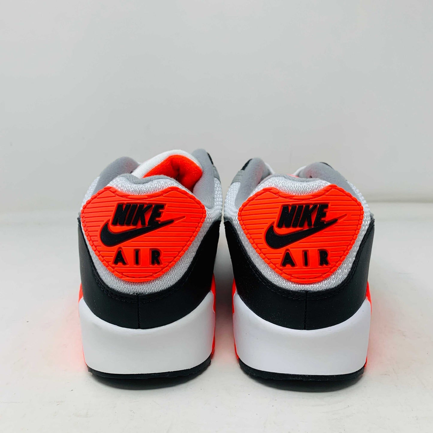 A pair of Nike Air Max 90 Infrared (2020) sneakers in gray, white, and bright orange sits on a pristine shoebox. Beside the shoes are stylish accessories: an orange keychain and spare orange soles.