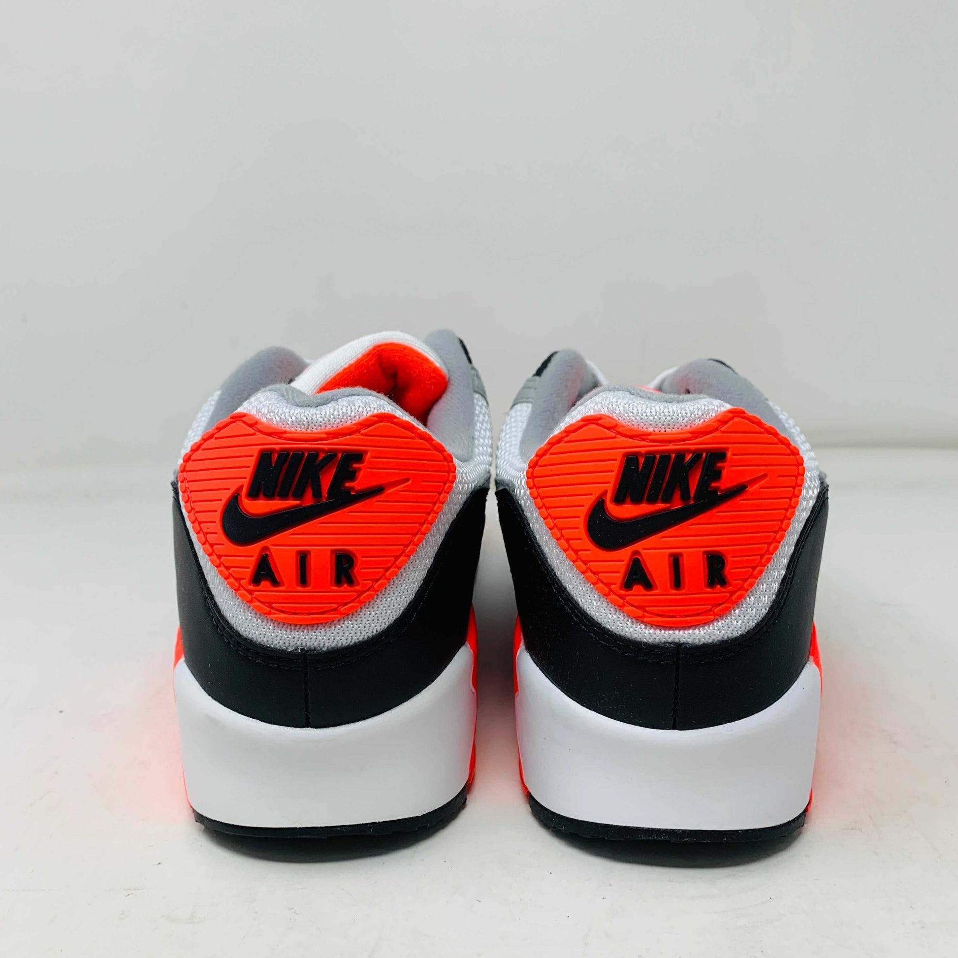 A pair of Nike Air Max 90 Infrared (2020) sneakers in gray, white, and bright orange sits on a pristine shoebox. Beside the shoes are stylish accessories: an orange keychain and spare orange soles.