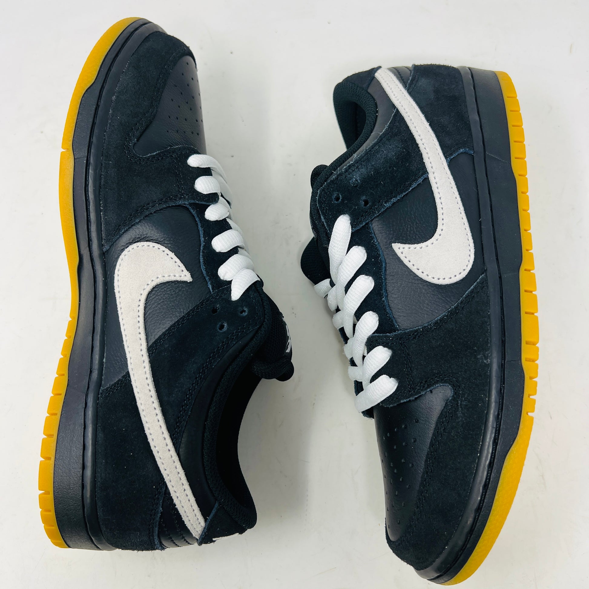 A black Nike Dunk Low Pro SB Black White Gum sneaker with white laces and swoosh sits on a purple Nike SB box. The 2025 shoe has a yellow sole, leather-texture finish, and both are in new condition.
