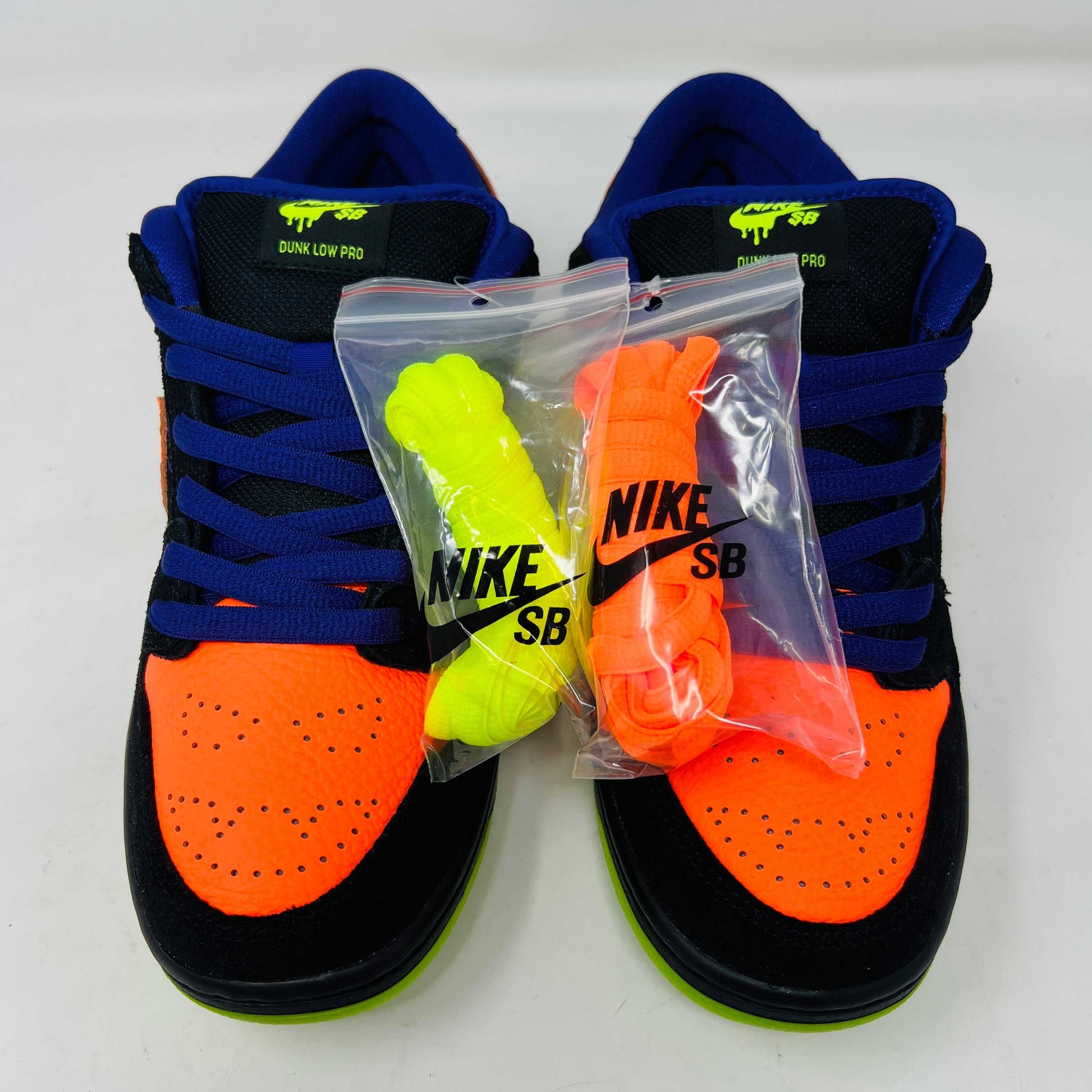 The Nike SB Dunk Low Night of Mischief sneakers feature Halloween-themed designs with orange swooshes, green soles, and spider web details. They come in a coffin-shaped box adorned with spooky jack-o-lanterns and webs, resting on a wooden floor.