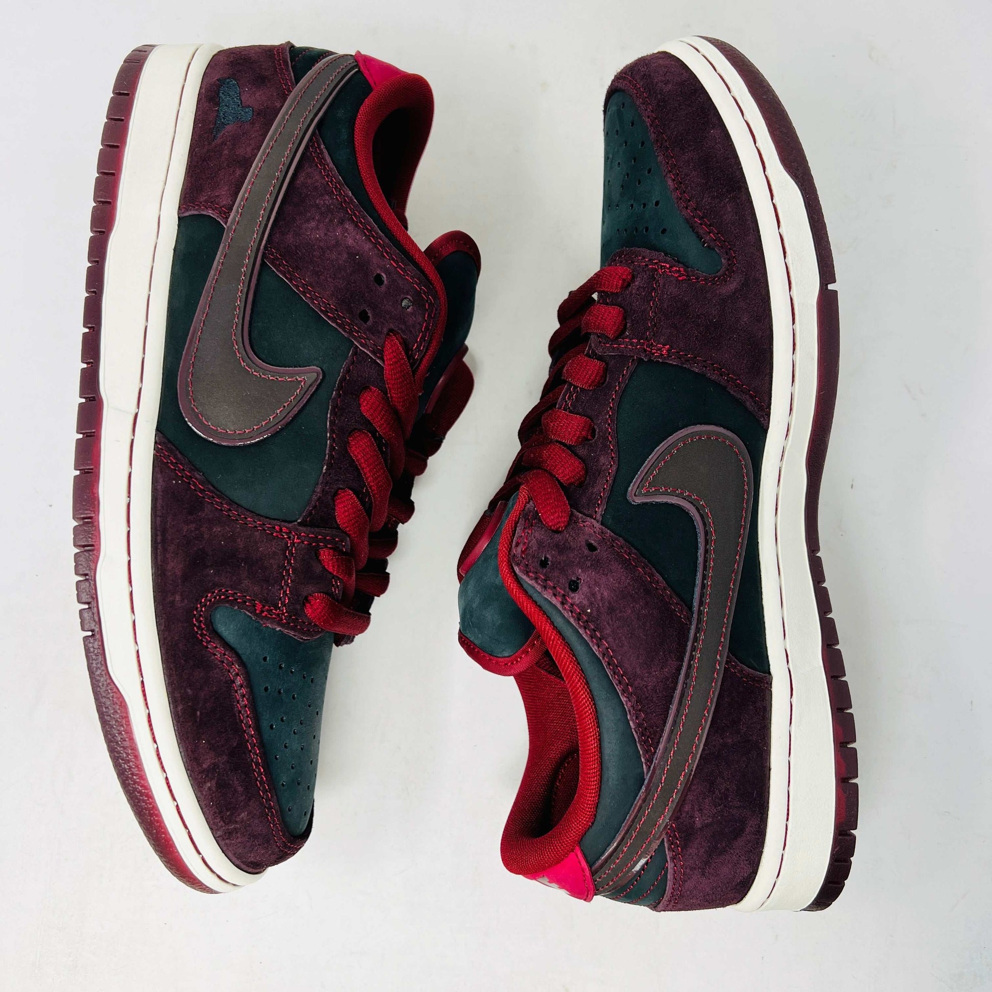 A BRAND NEW Nike SB Dunk Low Riot Skateshop sneaker featuring a black design with maroon accents, including a swoosh, laces, and a black sole with a white midsole, is displayed on top of its original white Nike SB shoebox.