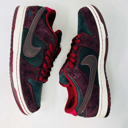 A BRAND NEW Nike SB Dunk Low Riot Skateshop sneaker featuring a black design with maroon accents, including a swoosh, laces, and a black sole with a white midsole, is displayed on top of its original white Nike SB shoebox.