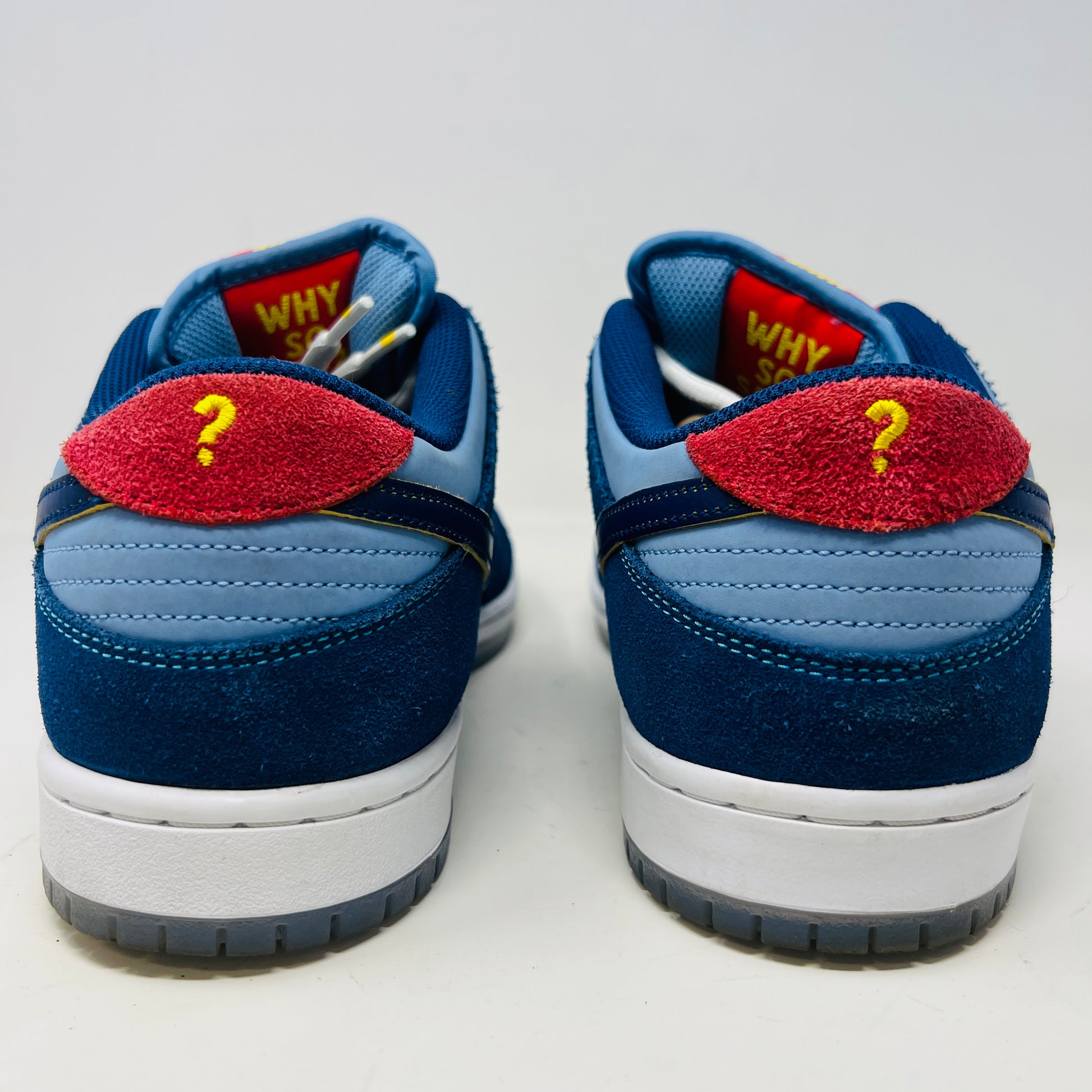 A Nike SB Dunk Low Pro Why So Sad? shoe, with blue uppers and white sole, rests on a purple box. It features an embroidered graphic on the heel, a dark blue swoosh, and extra laces in front. Price tags from 2022 are visible beside it.