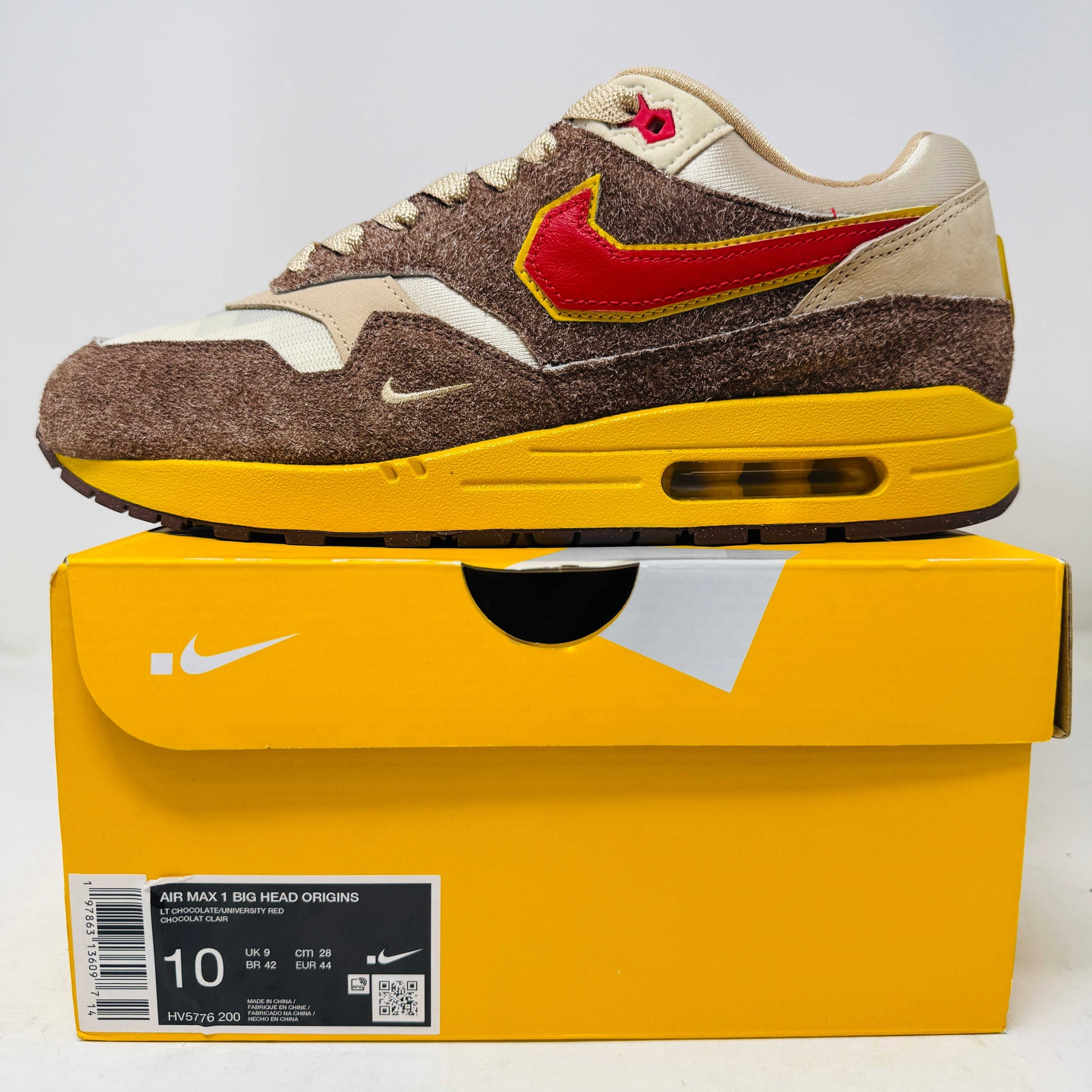 Nike Air Max 1 sneaker with brown suede, red and yellow swoosh, displayed on yellow Nike box
