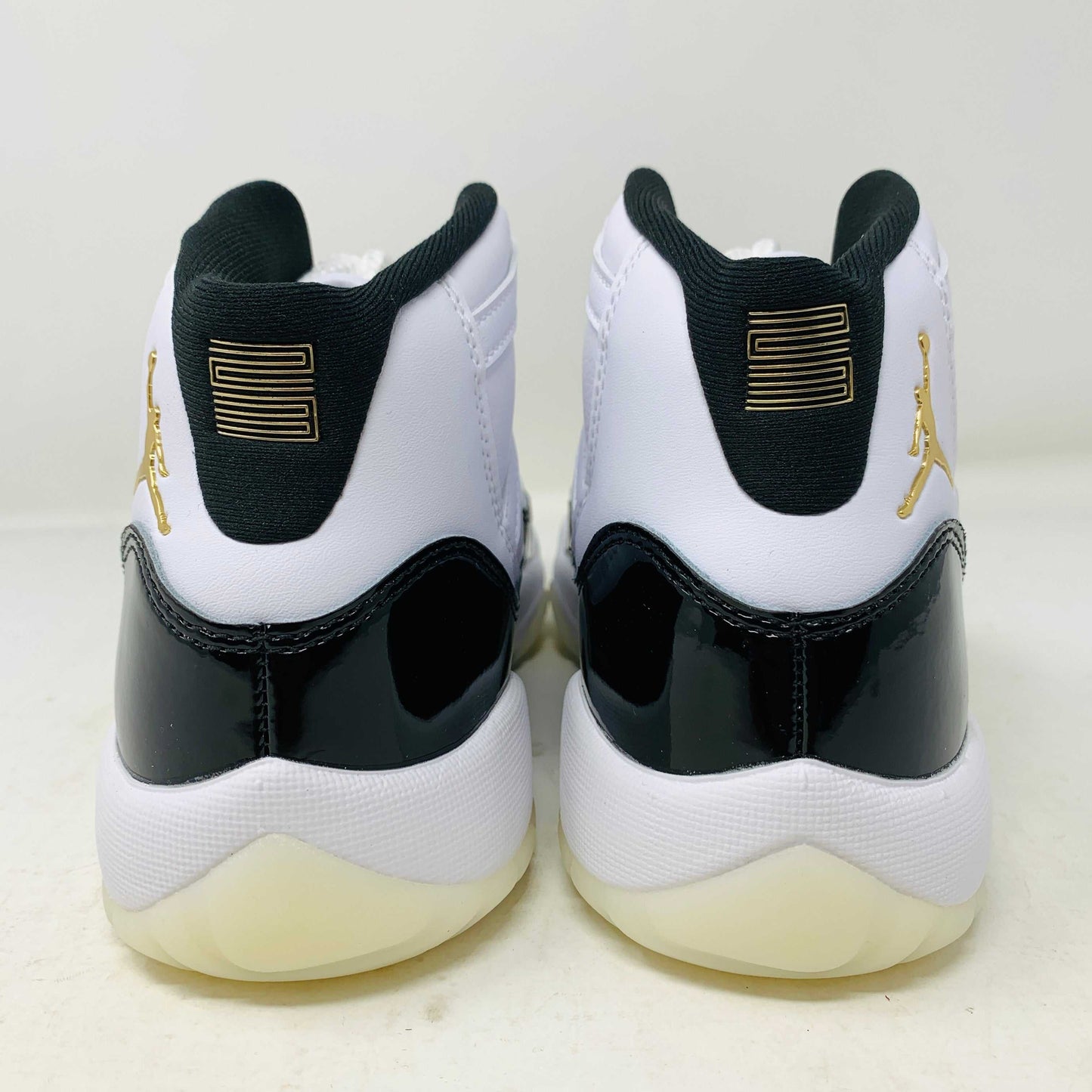 A pair of authentic Jordan 11 Retro DMP Gratitude (2023) (GS) sneakers, featuring a glossy white and black finish with a gold logo, is placed on top of a gray Nike Air shoebox.