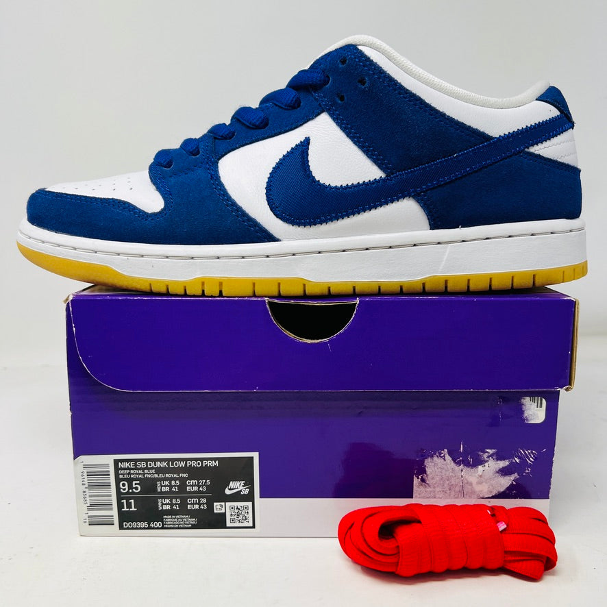The Nike SB Dunk Low Los Angeles Dodgers sneakers, featuring blue and white uppers with a white sole and yellow bottom, are showcased on a purple box. Red extra laces are neatly arranged in front, completing the look.